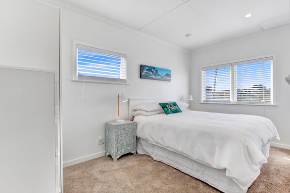 A bedroom with a bed, two bedside tables, a nightstand, and two windows with blinds. - The Little Blue Bach in Oneroa Village (Waiheke Island)