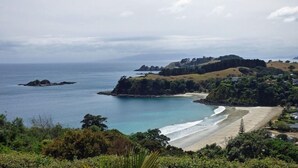 A coastal landscape with a beach, rocky outcrops, and a forested hill. - Ted's Cottage near Little Oneroa Beach (Waiheke Island)