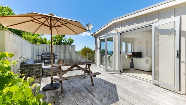 Um deck de madeira com uma mesa de piquenique e guarda-sol, uma churrasqueira e um espaço envidraçado com uma televisão.
