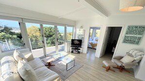Uma sala de estar ampla com um sofá grande, uma mesa de centro e uma televisão. Janelas grandes oferecem vista para o exterior.