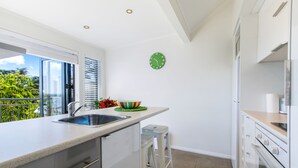 A modern kitchen with a large window, a sink, a colorful bowl on the counter, and a green clock on the wall. - Villa Casita at Palm Beach (Waiheke Island)