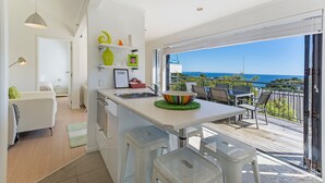 A modern kitchen with a view of the sea, a dining area with outdoor seating, and a balcony with a railing. - Villa Casita at Palm Beach (Waiheke Island)