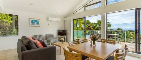 A modern living room with a sofa, TV, dining table, and chairs. Large windows offer a view of the outdoors.