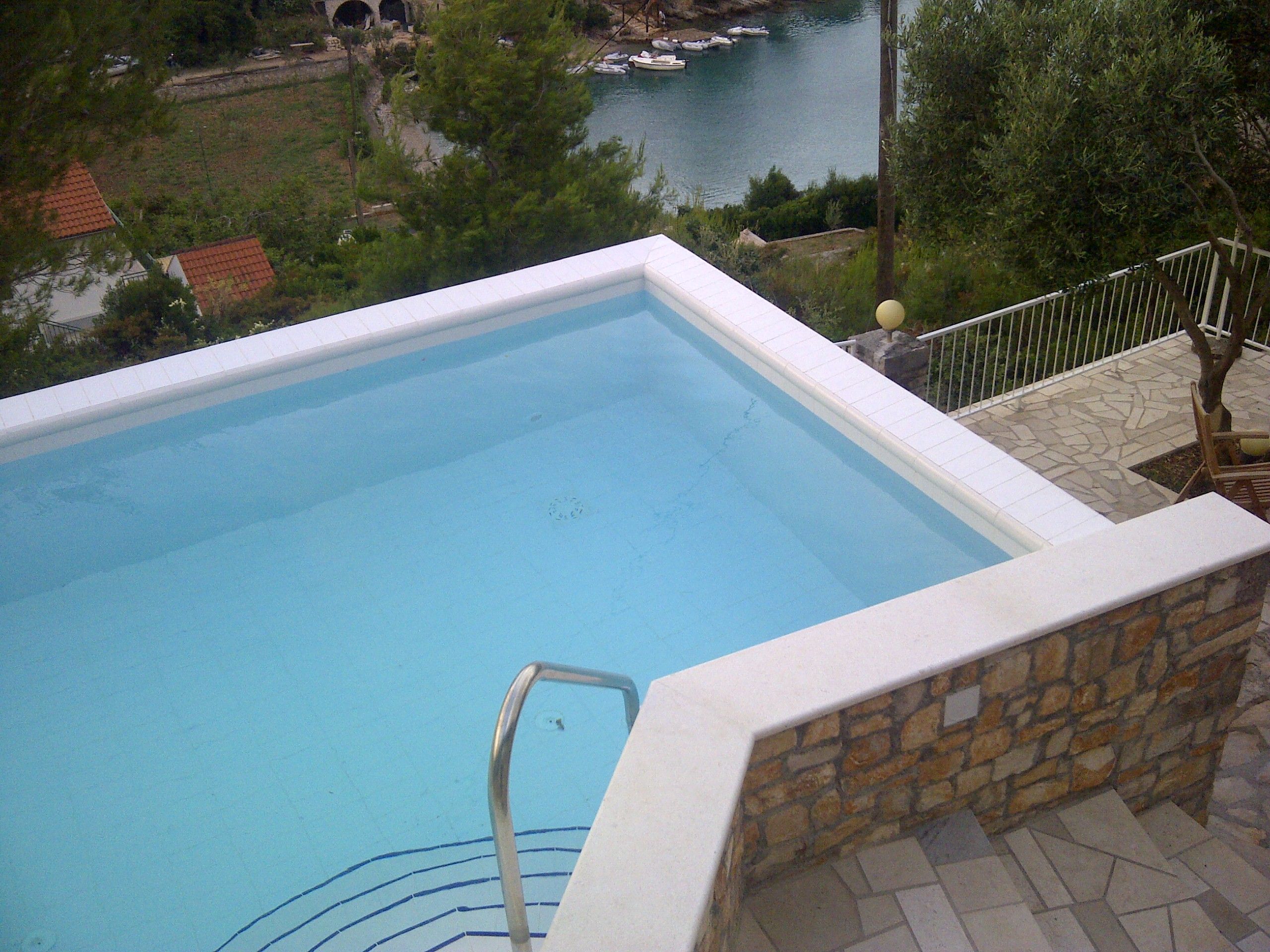 A swimming pool with a stone wall and metal railing, surrounded by a stone patio and a view of a body of water and greenery.