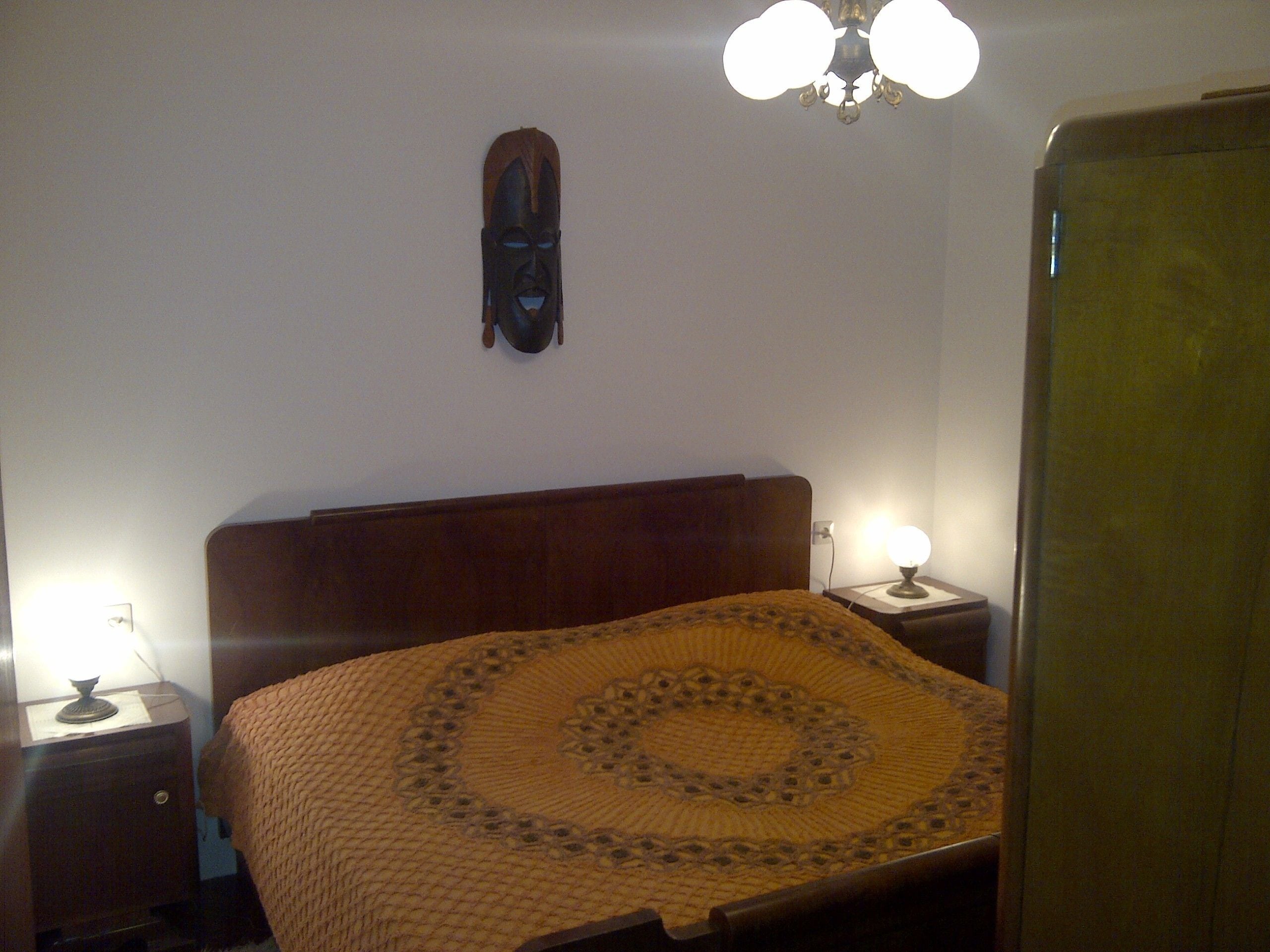 Une chambre à coucher avec une tête de lit en bois, une couvre-lit à motifs, deux lampes de chevet et un luminaire fixé au mur.