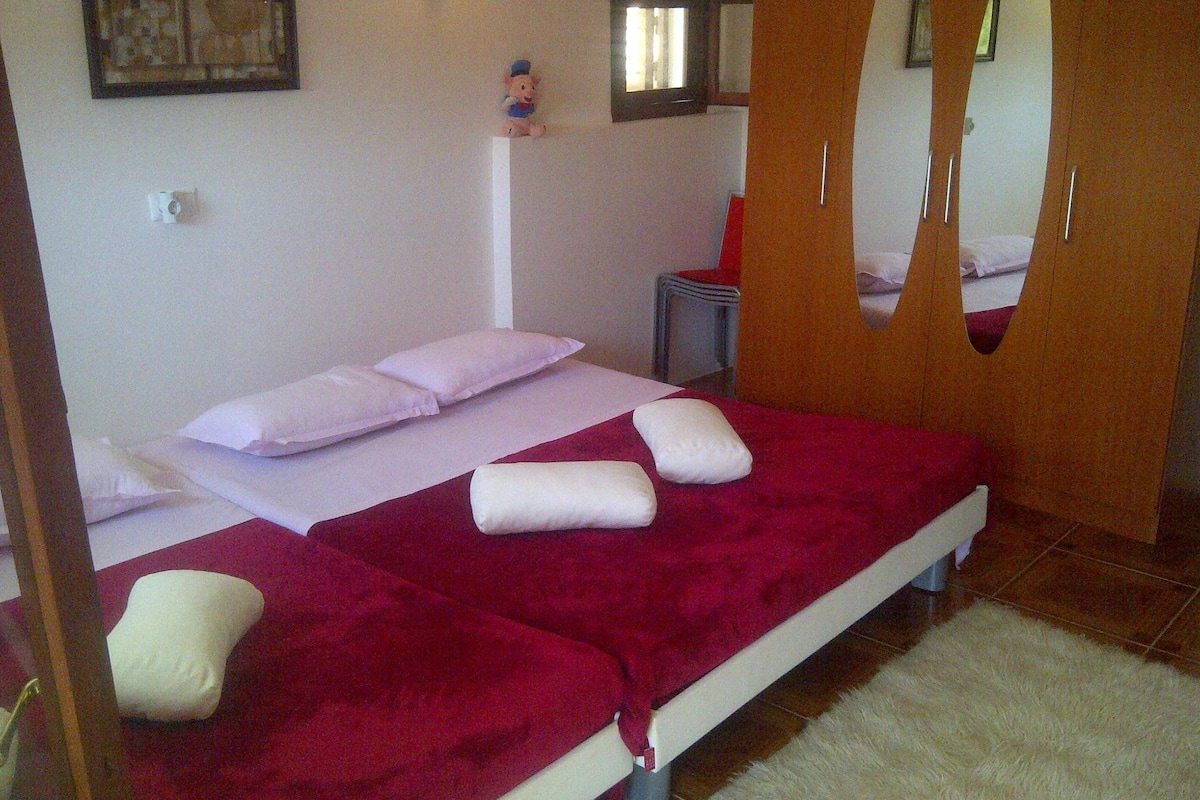A bedroom with a bed, pillows, a red blanket, a small table, and a mirror.