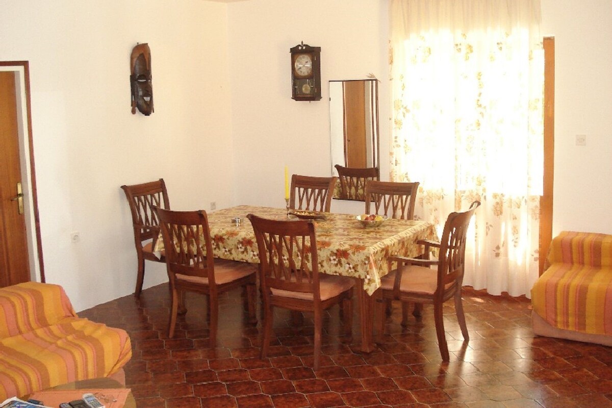 A dining area with a round table, six wooden chairs, a floral tablecloth, a clock on the wall, and a window with curtains.