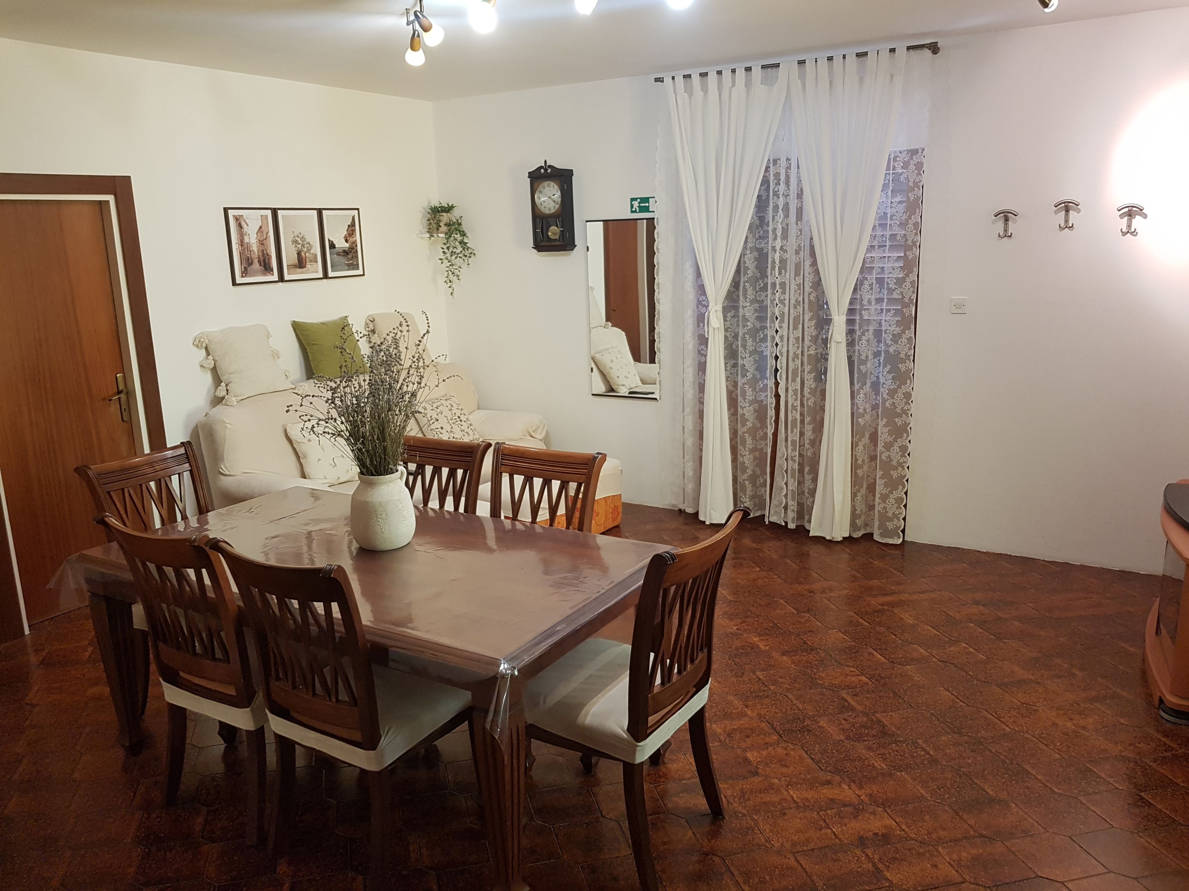 Un comedor con una mesa y sillas de madera, un jarrón con flores, un reloj en la pared y una ventana con cortinas.