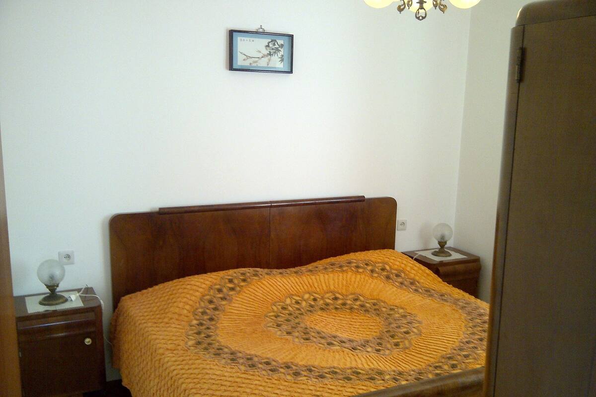 A bedroom with a wooden bed, a patterned blanket, a nightstand with a lamp, and a framed picture on the wall.