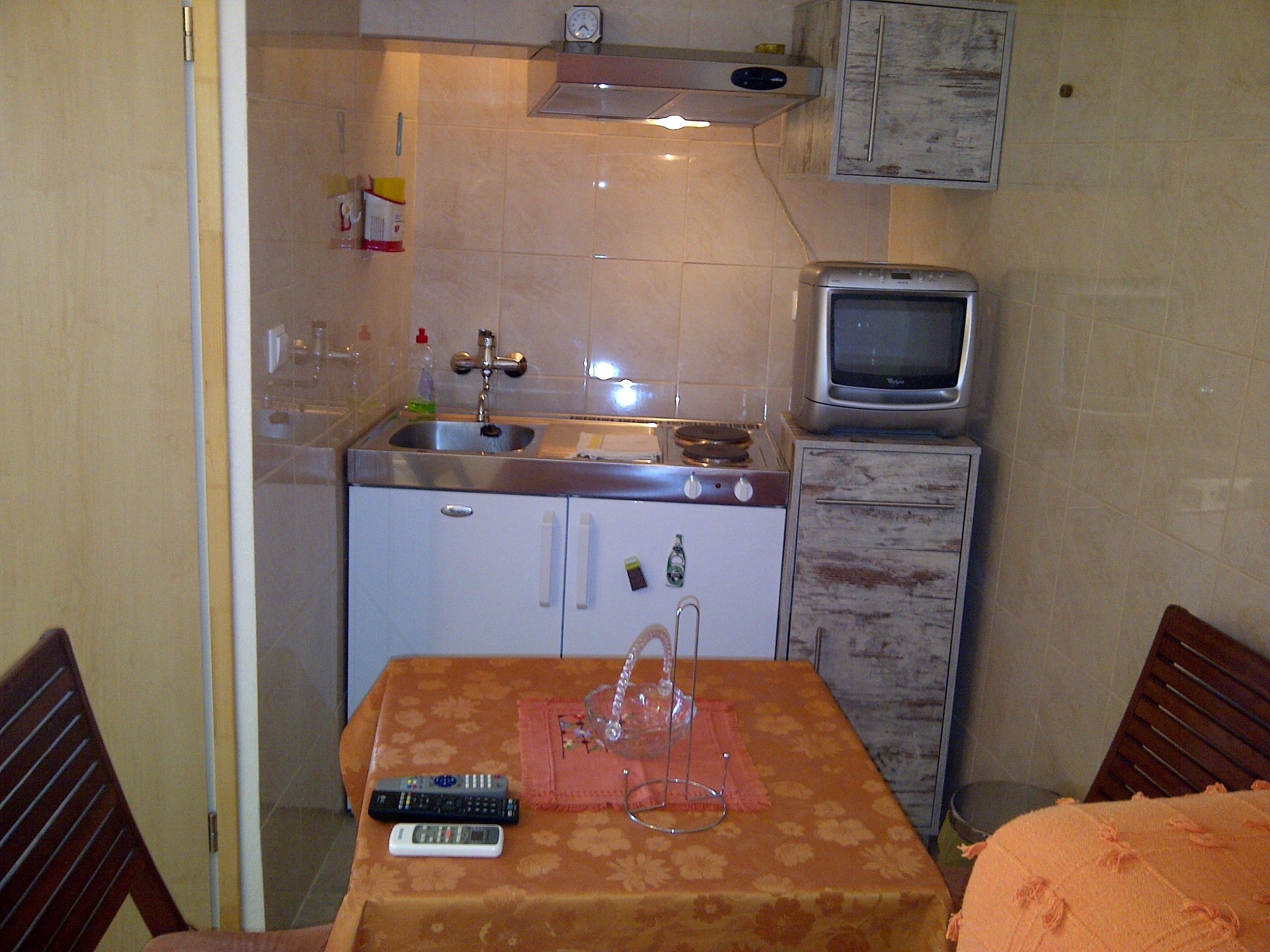 Cocina compacta con pileta, estufa y un refrigerador pequeño. Hay una mesa con un control remoto y un vaso encima.