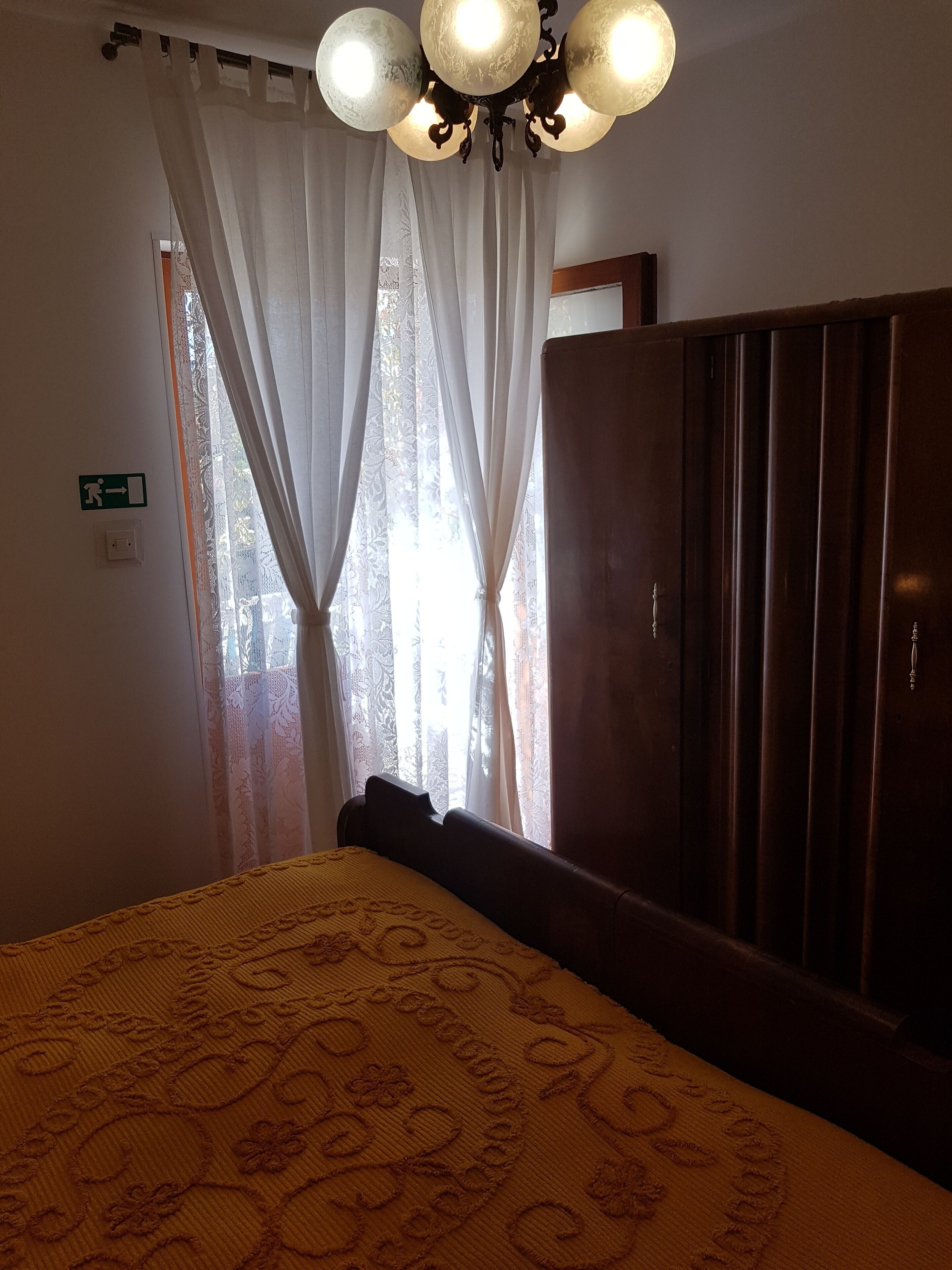 Een kamer met een bed, een houten garderobe, een kroonluchter en een raam met dunne gordijnen.