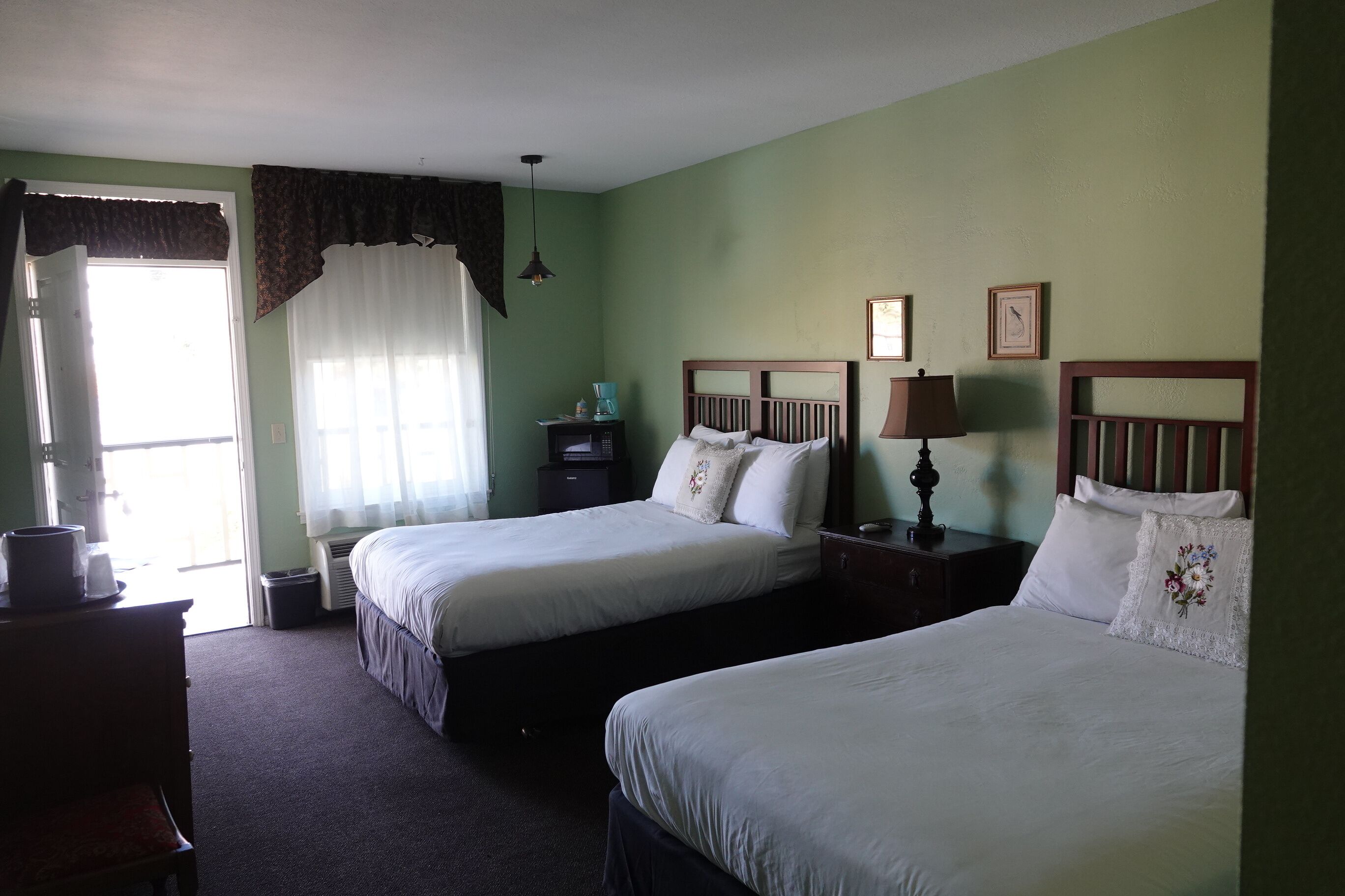 A hotel room with two single beds, a dresser, a desk with a lamp, and a window with curtains.