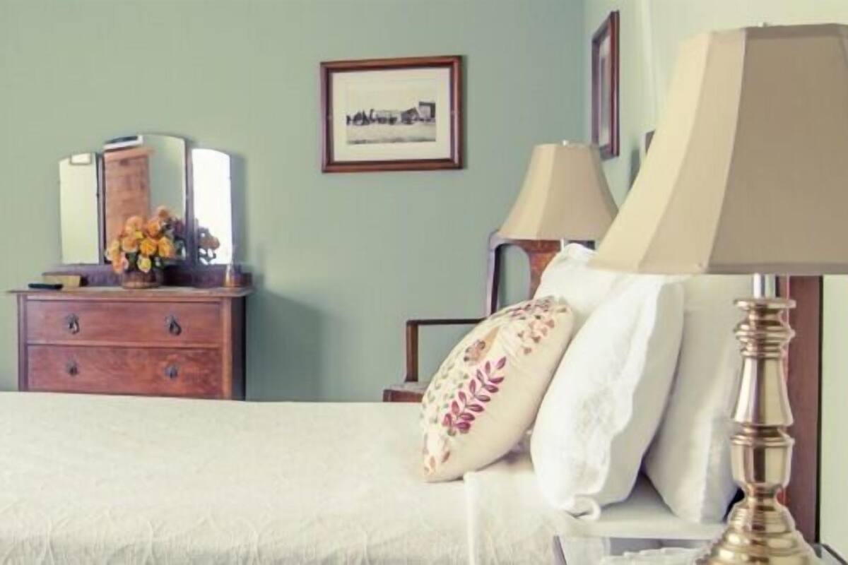 Una cama bien hecha con edredón blanco y almohadas decorativas, una mesita de noche de madera con un jarrón de flores y un cuadro en la pared.