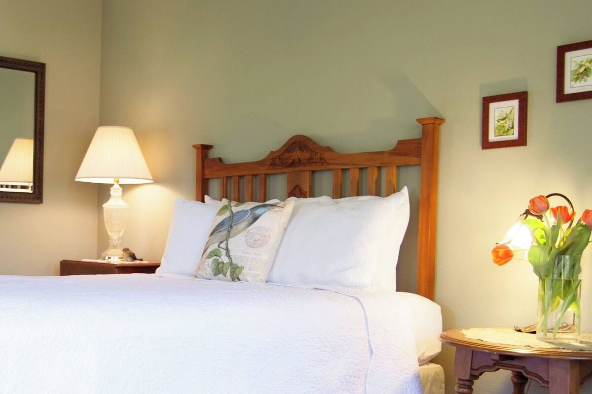 Una cama bien tendida con sábanas blancas, un cabecero de madera, una lámpara de noche y una mesita con un jarrón de tulipanes.