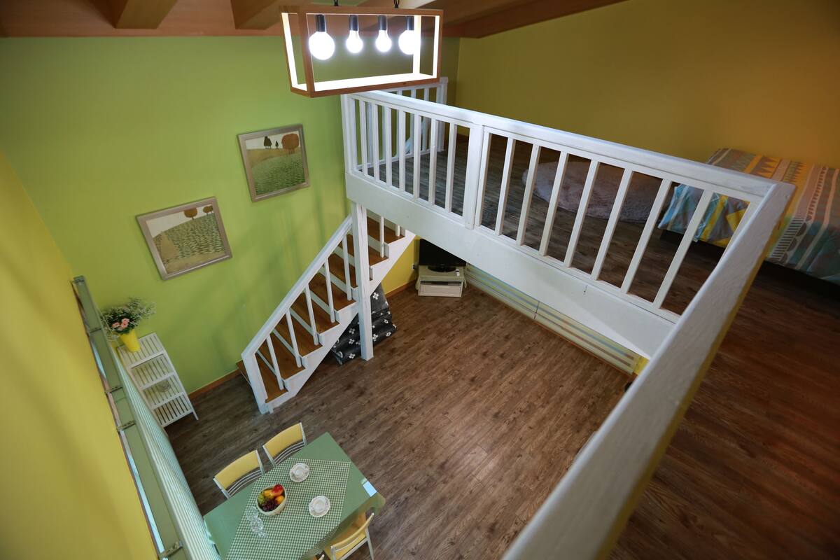 A loft bedroom with a wooden floor, a green table with a fruit bowl, and a white railing.
