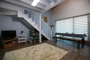 A living room with a staircase, a flat-screen TV, a bench, and a window with blinds.
