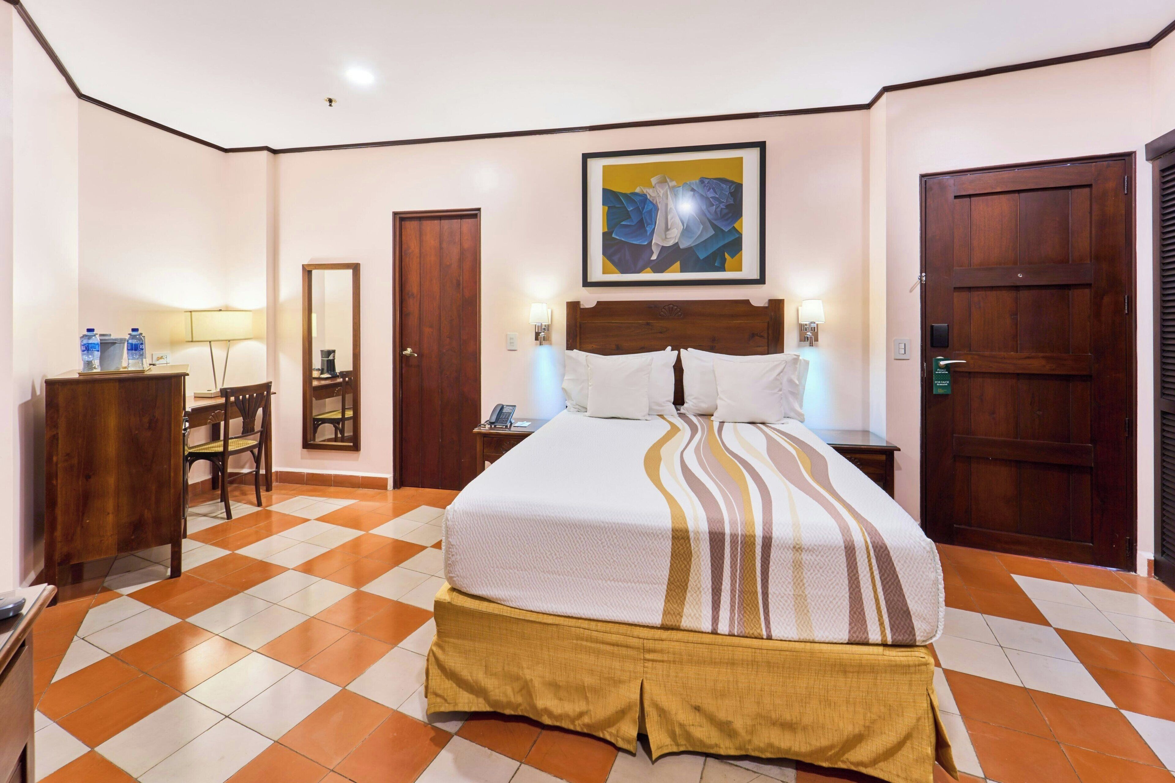 A hotel room with a checkered floor, a bed with a striped bedspread, a wooden headboard, a desk with a chair, a mirror, and a door.