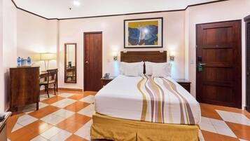 A hotel room with a checkered floor, a bed with a striped bedspread, a wooden headboard, a desk with a chair, a mirror, and a door.