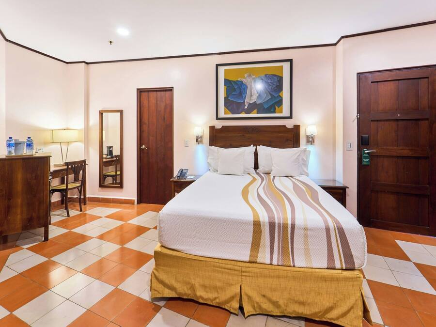 A hotel room with a checkered floor, a bed with a striped bedspread, a wooden headboard, a desk with a chair, a mirror, and a door.