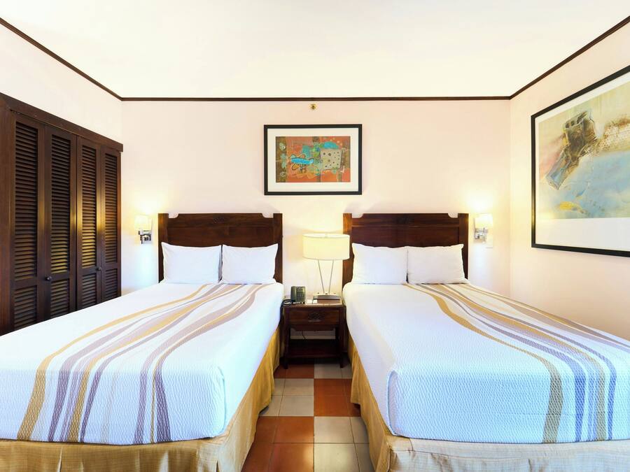 A hotel room with two beds, a nightstand, a lamp, and artwork on the walls.