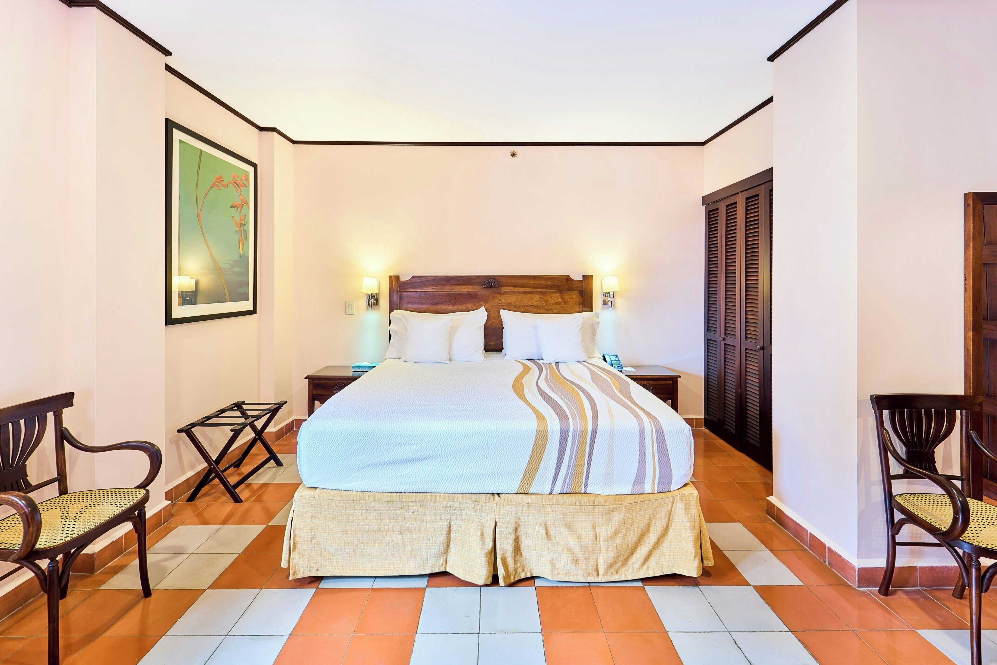 A hotel room with a bed, two bedside tables, two chairs, a painting on the wall, and a tiled floor.