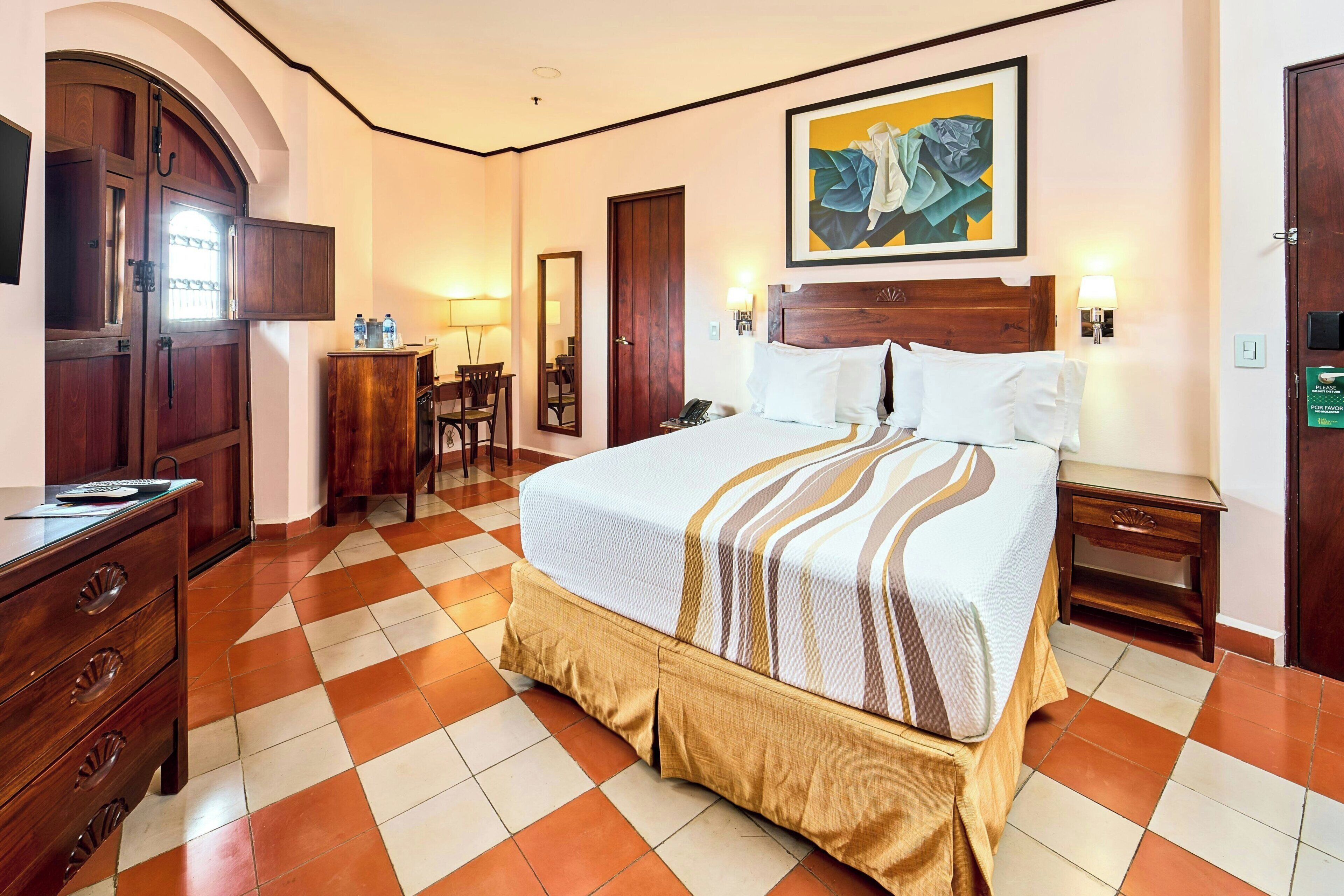 A hotel room with a bed, a desk, a chair, a wardrobe, and a painting on the wall.