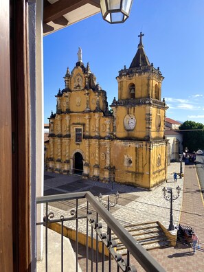 Una iglesia amarilla histórica con un campanario y una estatua en la parte superior, vista desde un balcón con barandilla de metal y un farol abajo.