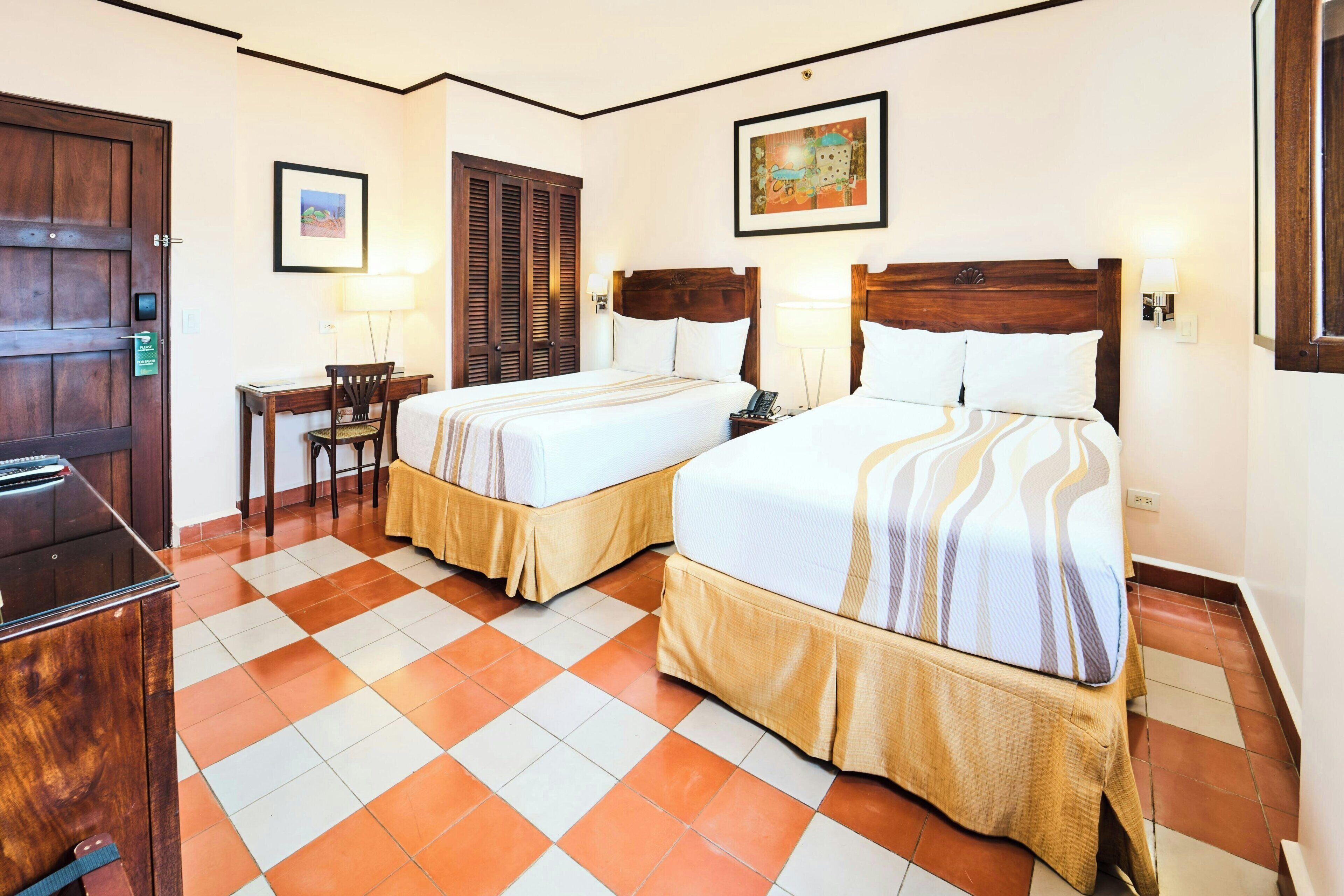 A hotel room with two beds, a desk, a chair, and a door.