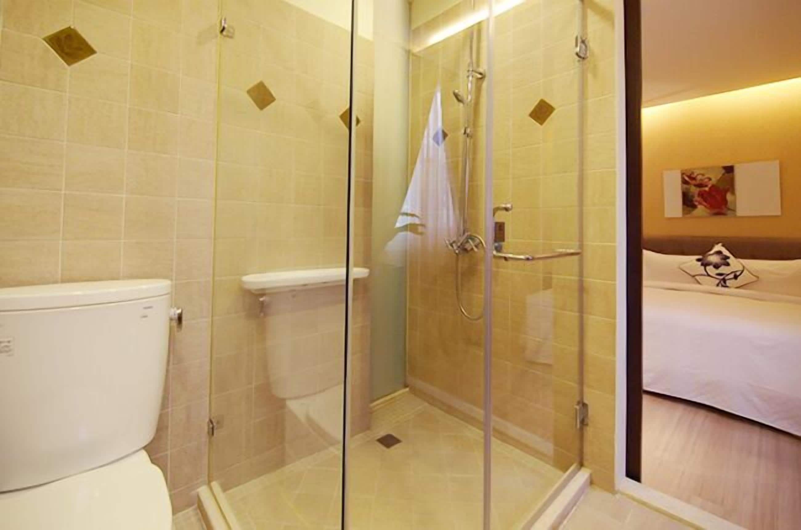 A hotel bathroom with a glass-enclosed shower, a toilet, and a bed visible through a glass partition.