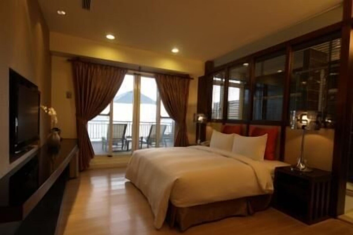 A hotel room with a large bed, a television, a balcony with a view, and a desk with a lamp.
