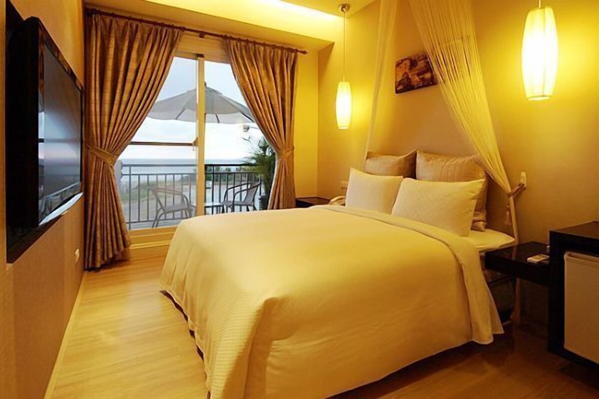A hotel room with a large bed, a TV, a desk, and a balcony with curtains.