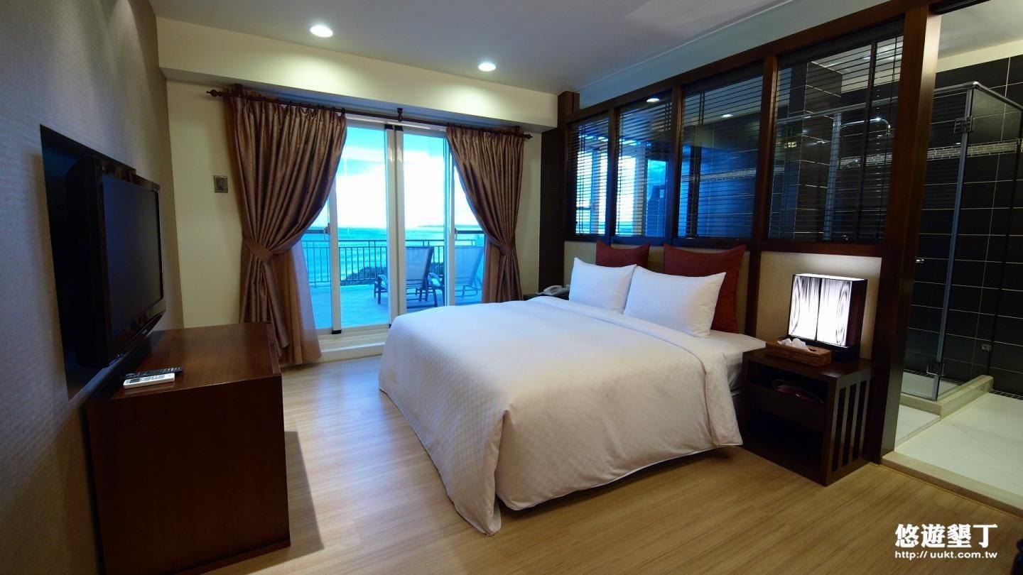 A hotel room with a large bed, a TV on a wooden cabinet, a bedside table with a lamp, and a balcony with curtains.