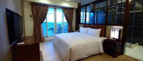 A hotel room with a large bed, a TV on a wooden cabinet, a bedside table with a lamp, and a balcony with curtains.