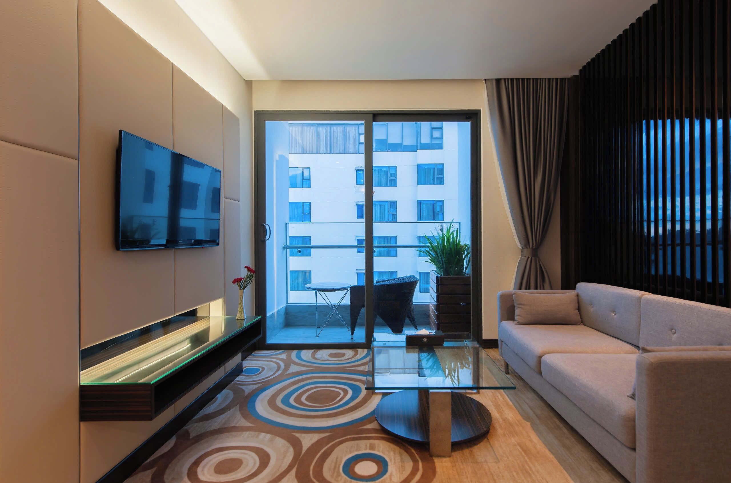Un salon moderne avec un téléviseur à écran plat fixé au mur, une table basse en verre, un canapé beige et un tapis à motifs.
