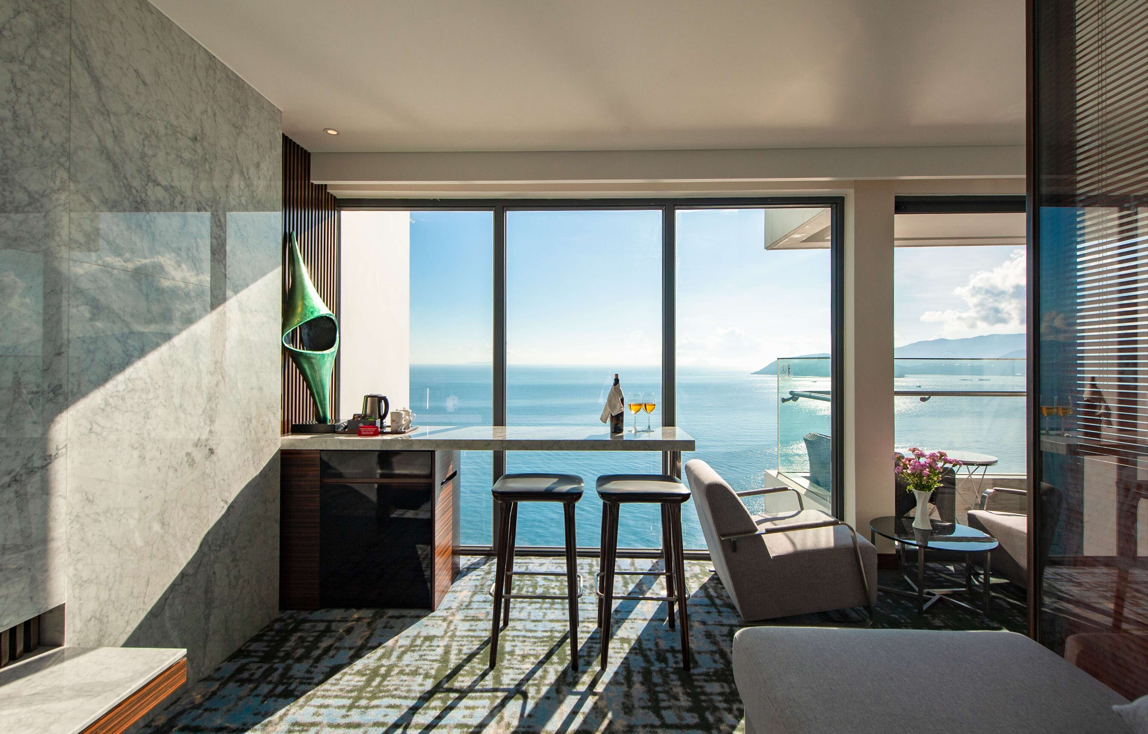 Une chambre d’hôtel moderne dotée d’une grande fenêtre donnant sur la mer. Elle comprend un coin repas avec une table et des chaises, un canapé et un petit balcon également équipé d’une table et de chaises.