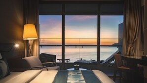 Habitación de hotel con un ventanal grande que da a una masa de agua, una cama, un escritorio y una silla.