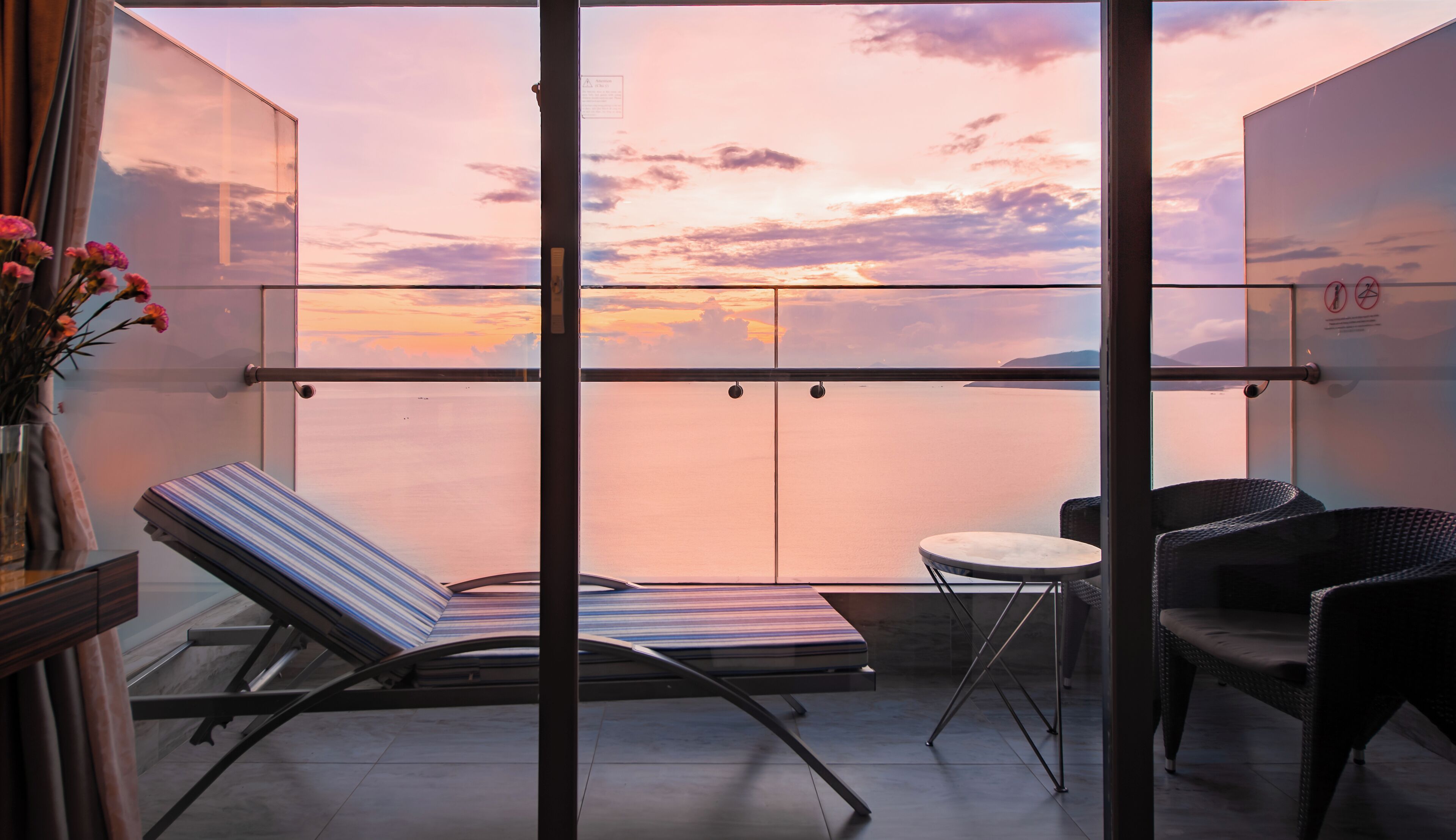 Ein Balkon mit Blick auf einen Sonnenuntergang über dem Wasser, ein gestreifter Liegestuhl, ein kleiner runder Tisch und Korbstühle.