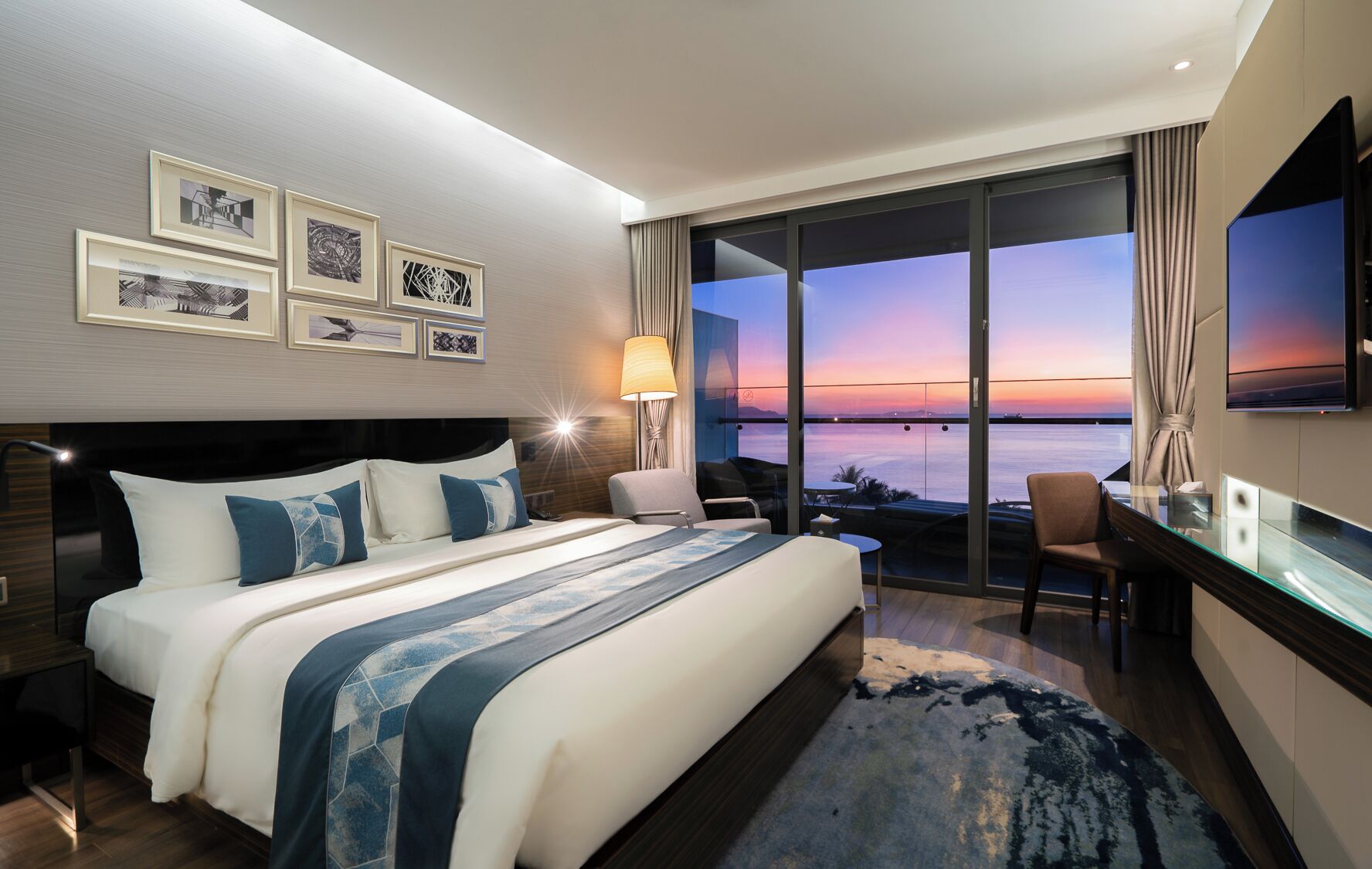 A modern hotel room with a large bed, a desk, and a view of the sunset over the water.