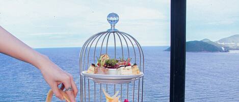 A table set with a tea kettle, cups, and a tiered dessert stand with various pastries and fruits, overlooking a body of water.