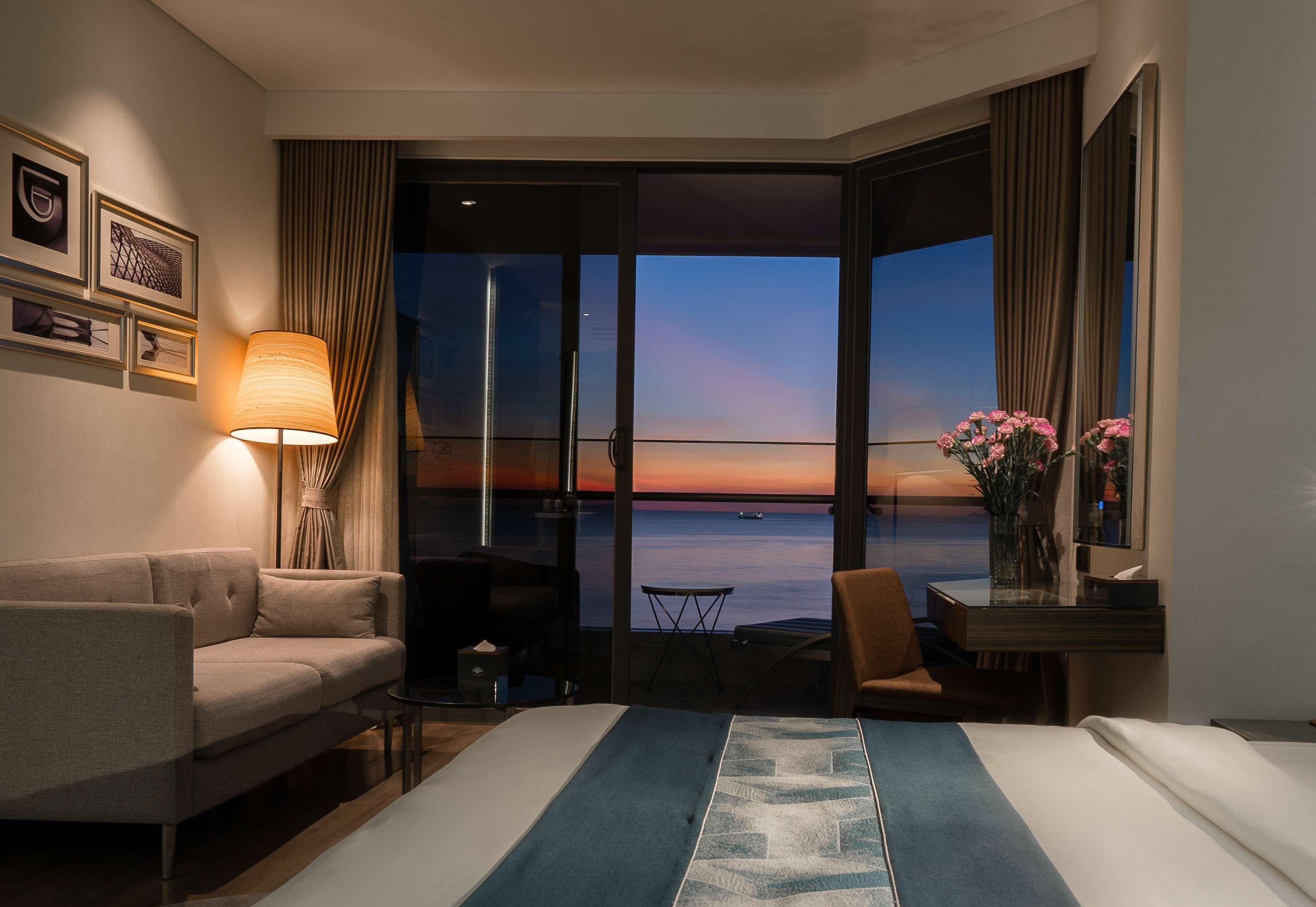 Ein Hotelzimmer mit Bett, einer Couch, einem Schreibtisch, einem Sessel, einer Lampe und Blick auf den Sonnenuntergang über dem Meer.