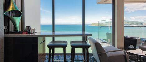 A modern room with a large window overlooking the sea, a bar area with stools, and a seating area with a sofa and chairs.