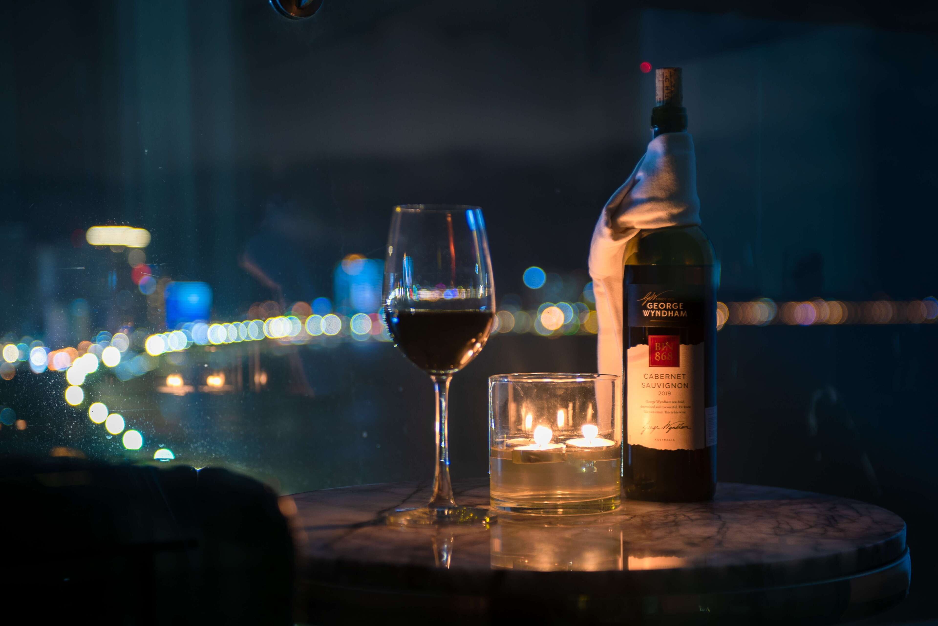 Une bouteille de George Wyndham Cabernet Sauvignon 2019, un verre de vin rouge et une bougie allumée sur une table, avec en arrière-plan une vue sur les lumières de la ville.