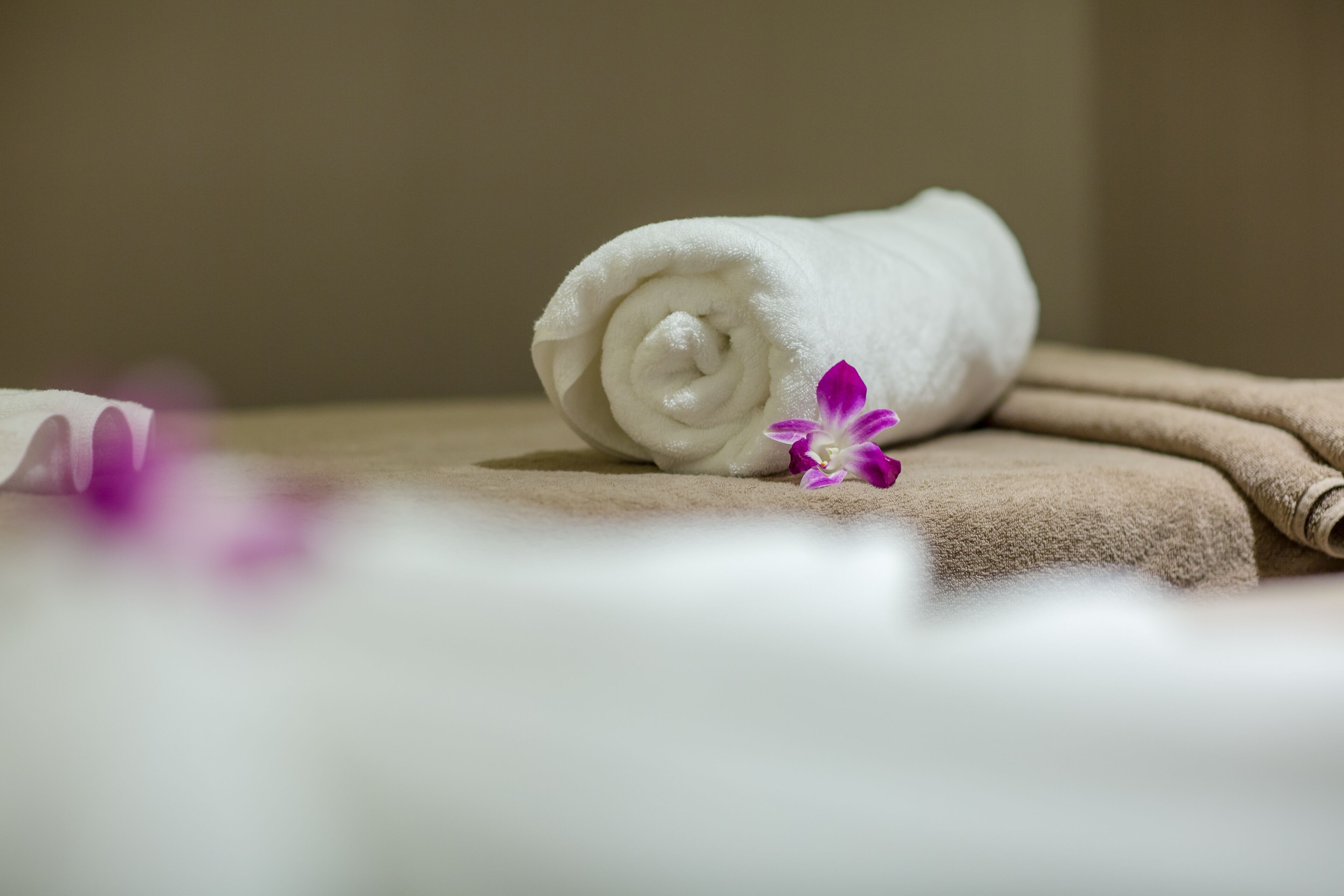 A rolled white towel and a purple flower on a beige surface.