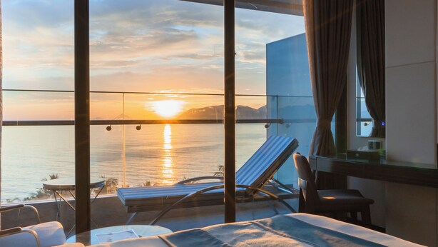 A balcony with a view of the sunset over the water, a lounge chair, and a bed with a view. - Queen Ann Nha Trang (Nha Trang)