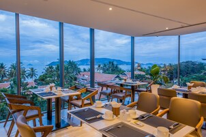 Ristorante con ampie vetrate che si affaccia su un paesaggio costiero. Tavoli apparecchiati con posateria e tovaglioli.