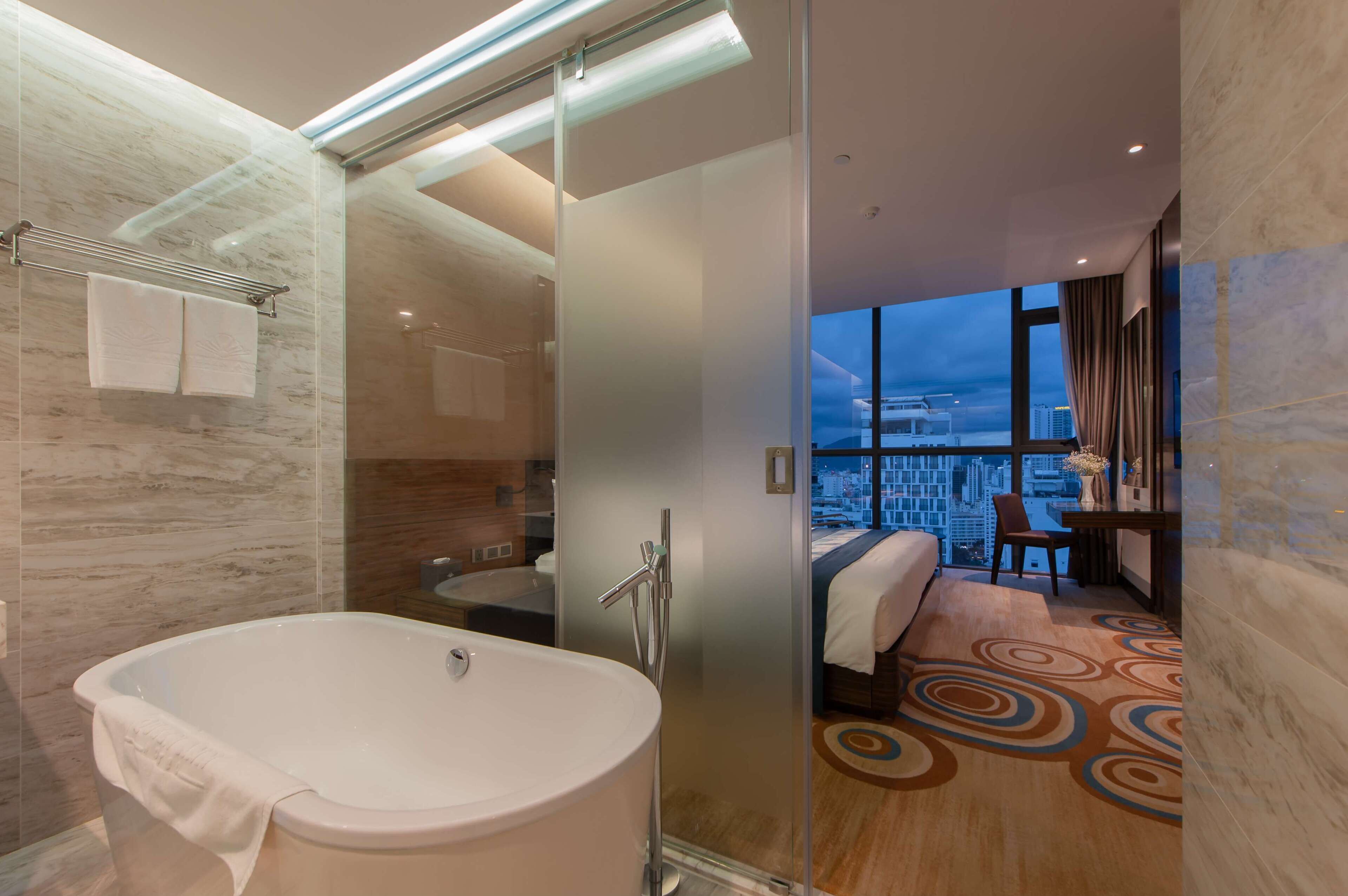 A modern hotel room with a glass-enclosed shower, a large bathtub, a bed with a view of the city, and a small desk area.