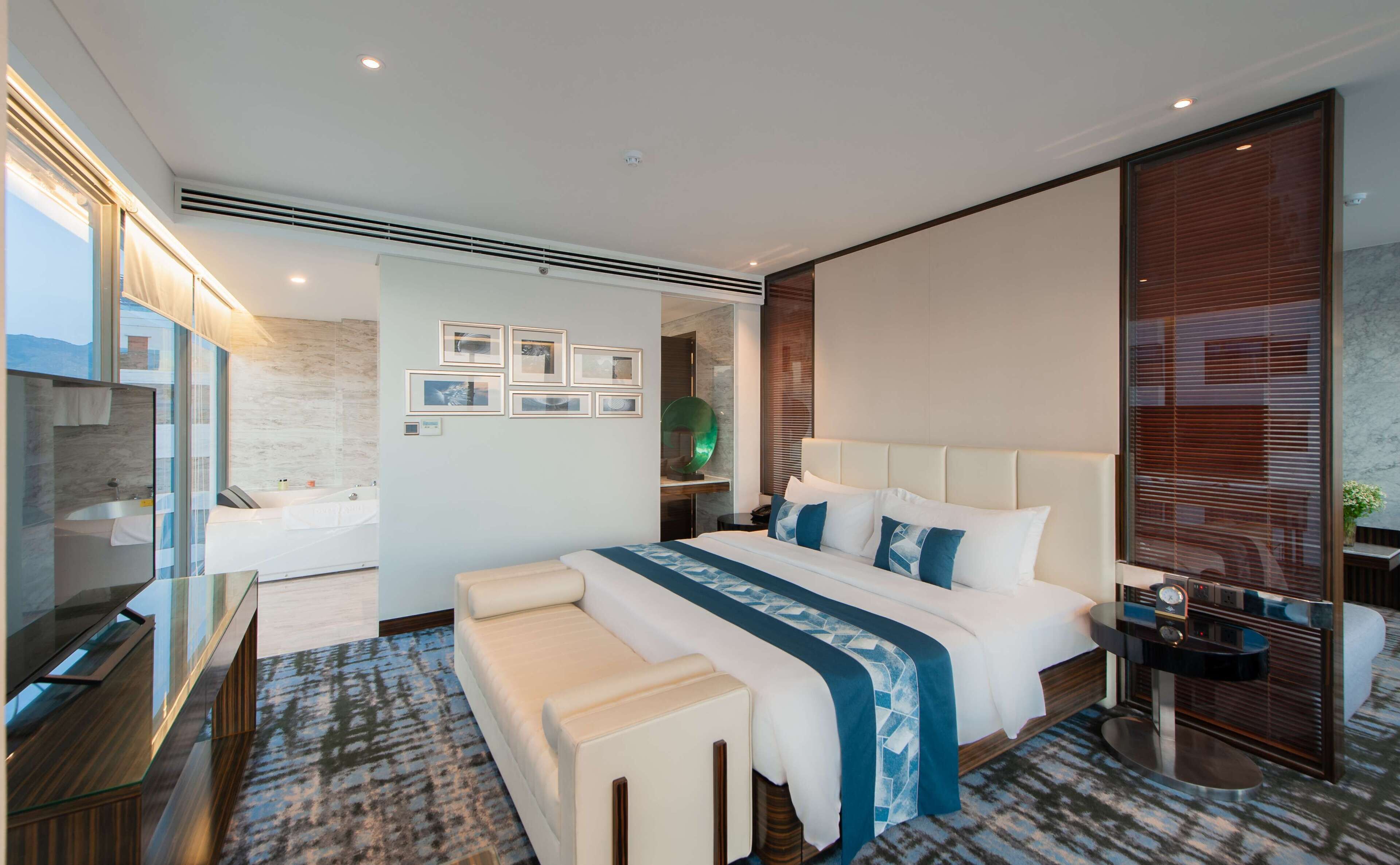 A modern hotel room with a large bed, a separate seating area, a glass-enclosed shower, and a view of the mountains.