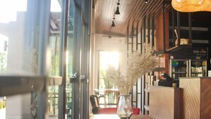 A restaurant interior with a wooden table, a vase with dried plants, and a view of the street outside. - Bunprasop Garden Hotel (Nakhon Si Thammarat)