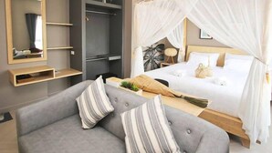 A hotel room with a grey sofa, a bed with a canopy, a small table with a plant, and a mirror on the wall. - Bunprasop Garden Hotel (Nakhon Si Thammarat)