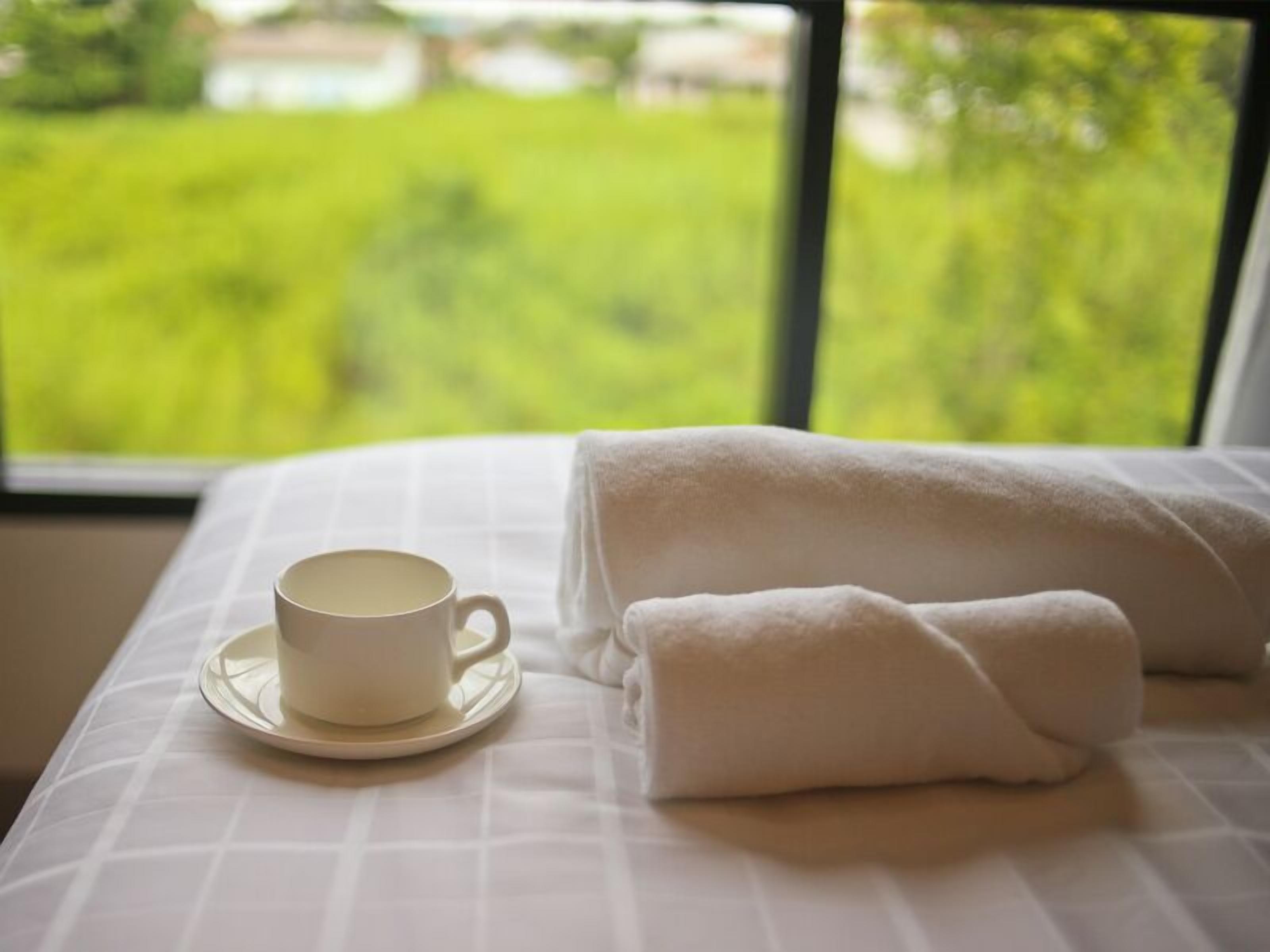 Una cama bien tendida con una taza y platillo blancos, una toalla doblada y vistas a un paisaje verde a través de una ventana.